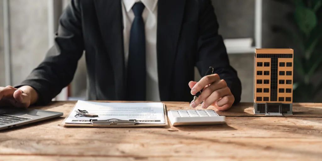 A financial advisor sitting at a desk performing calculations. He is wearing a dark colour suit and has a notepad open with a calculator under his left hand