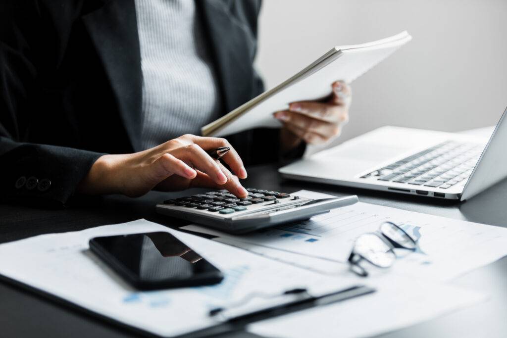 A female accountant is sitting at a desk in an office. She is wearing a business suit. She is looking down at a calculator and appears to be working on some calculations.