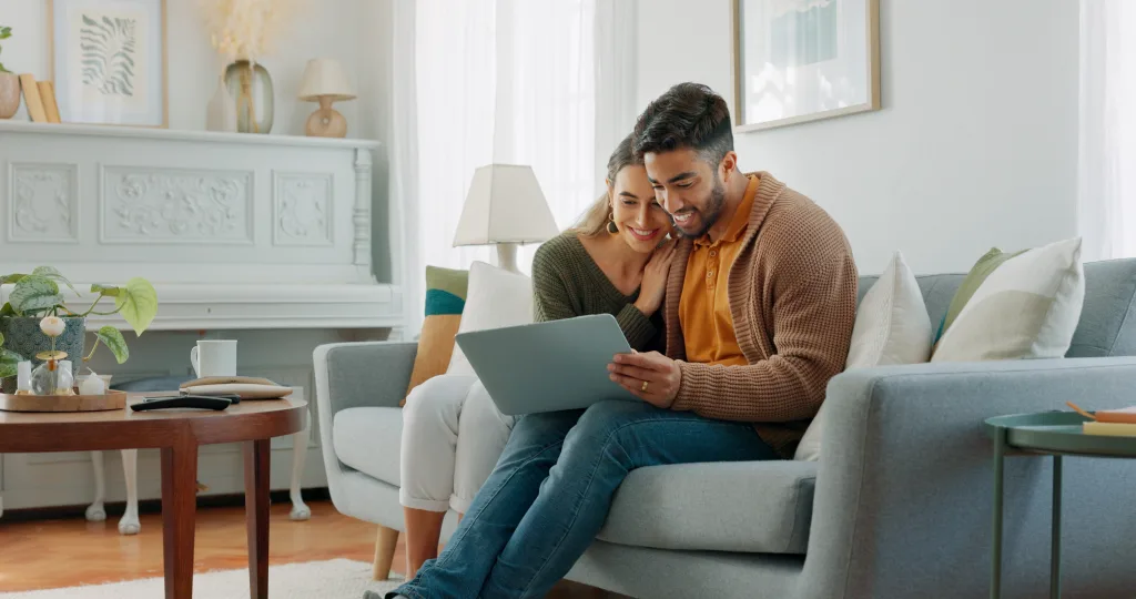 A man and a woman are sitting on a grey sofa with a laptop on the man's lap. They are both focused on the laptop and smiling